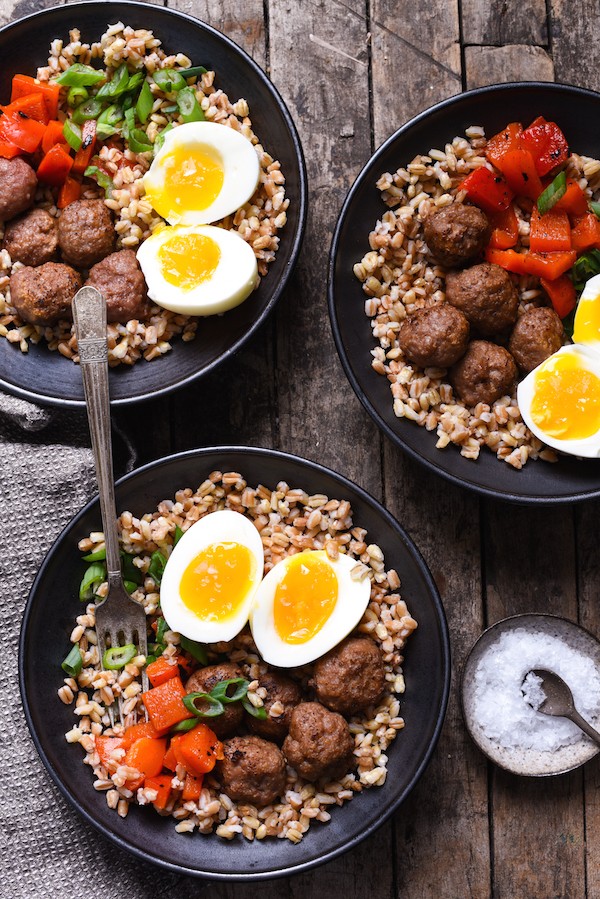 Overhead view of three grain bowls topped with sausage meatballs, fresh vegetables, and boiled egg.
